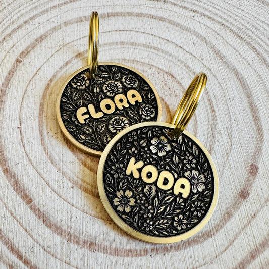 Two deep engraved black and gold dog tags with floral patterns on a wooden surface.