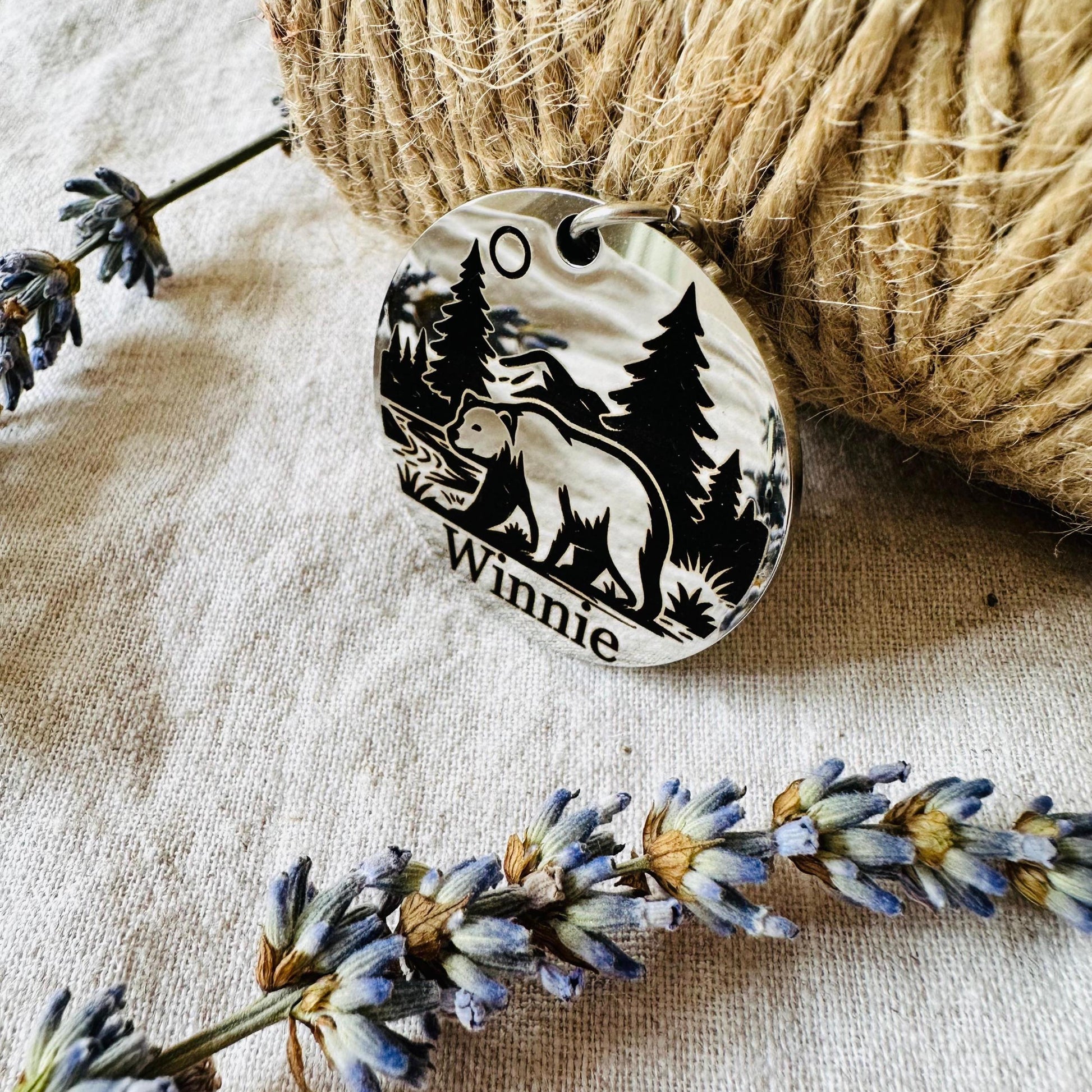 Round metal tag with bear design and 'Winnie' engraving on a textured fabric background with twine and lavender.