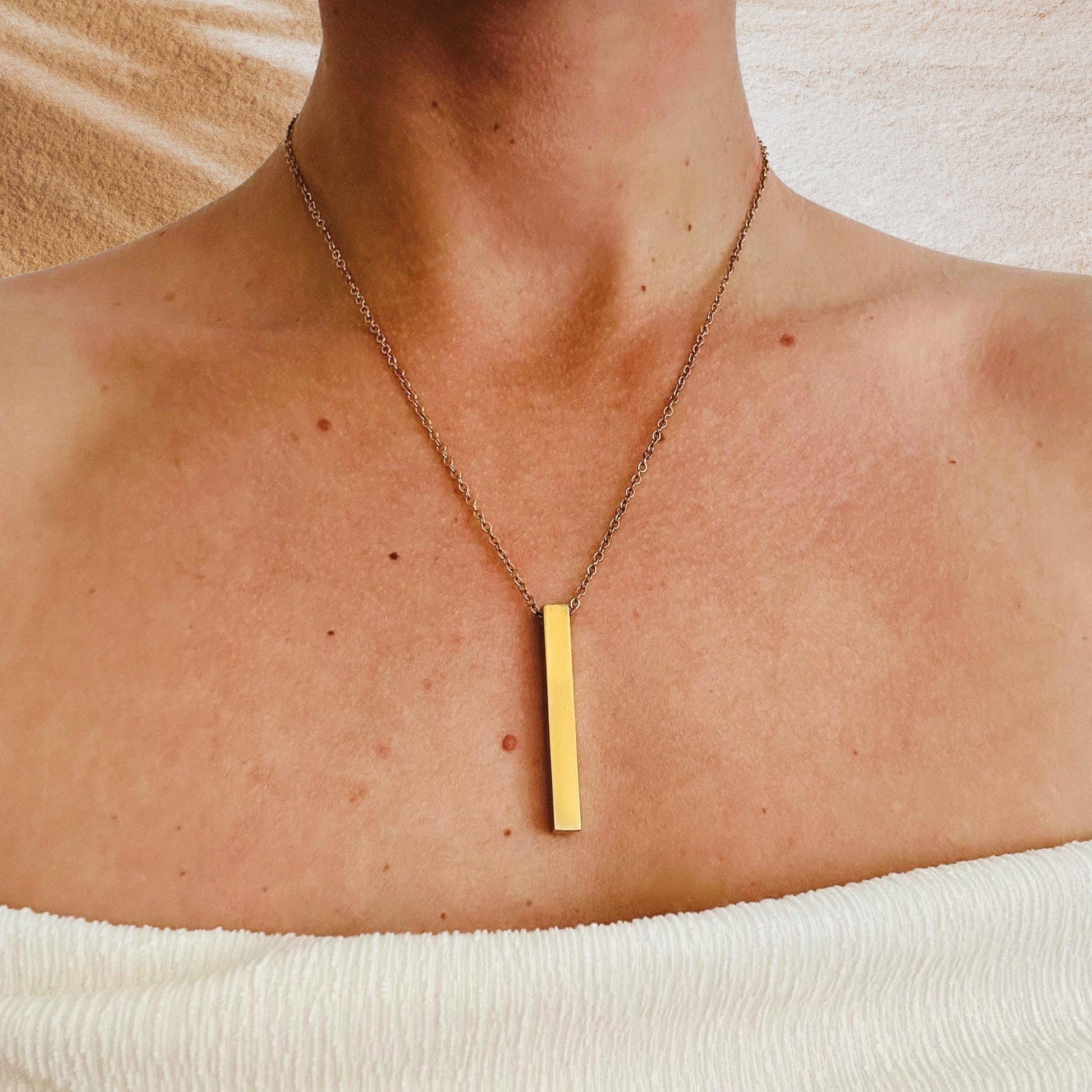 Gold necklace with a vertical bar pendant on a person's neck against a neutral background