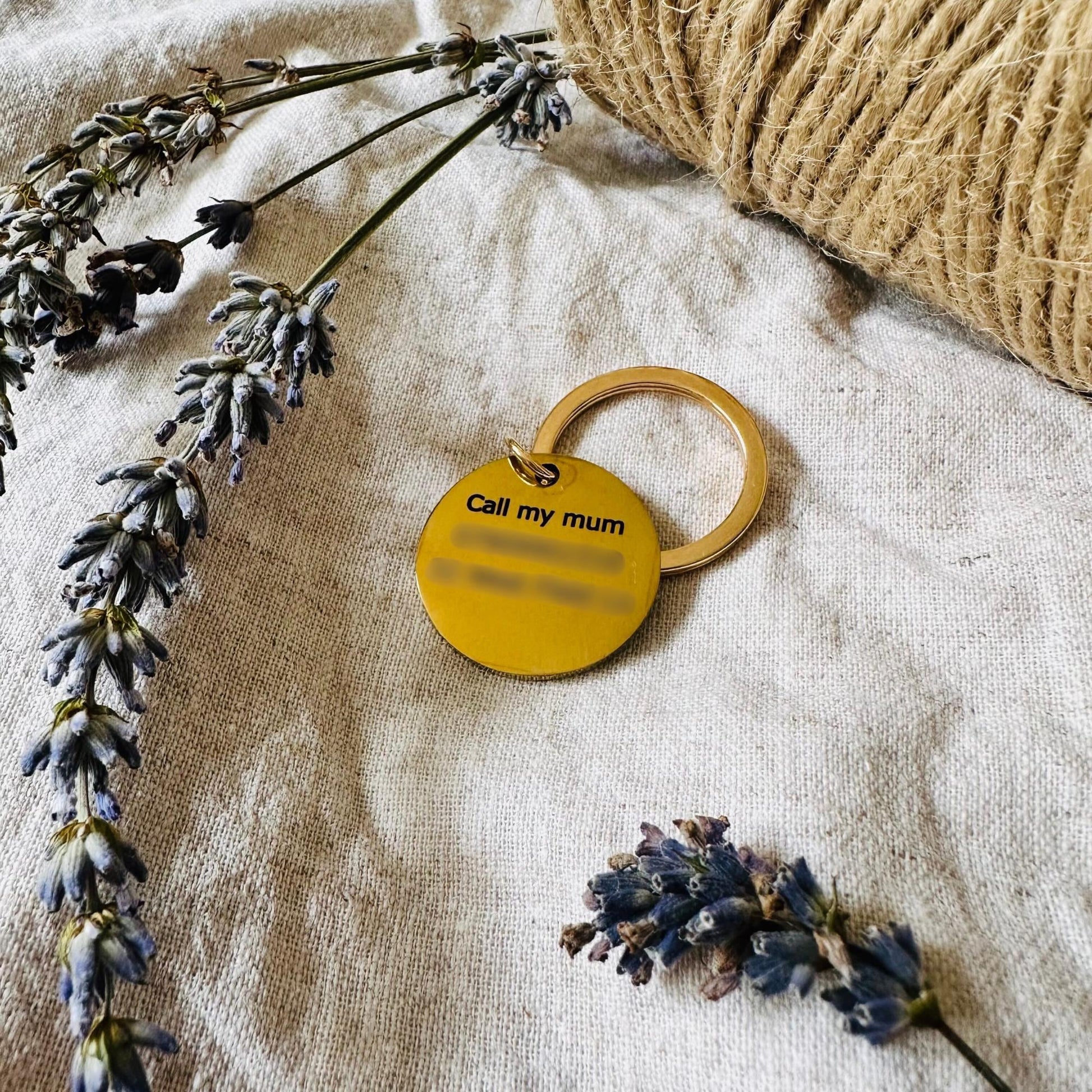 gold dog tag with 'Call my mum' text on a textured surface with lavender flowers and twine.