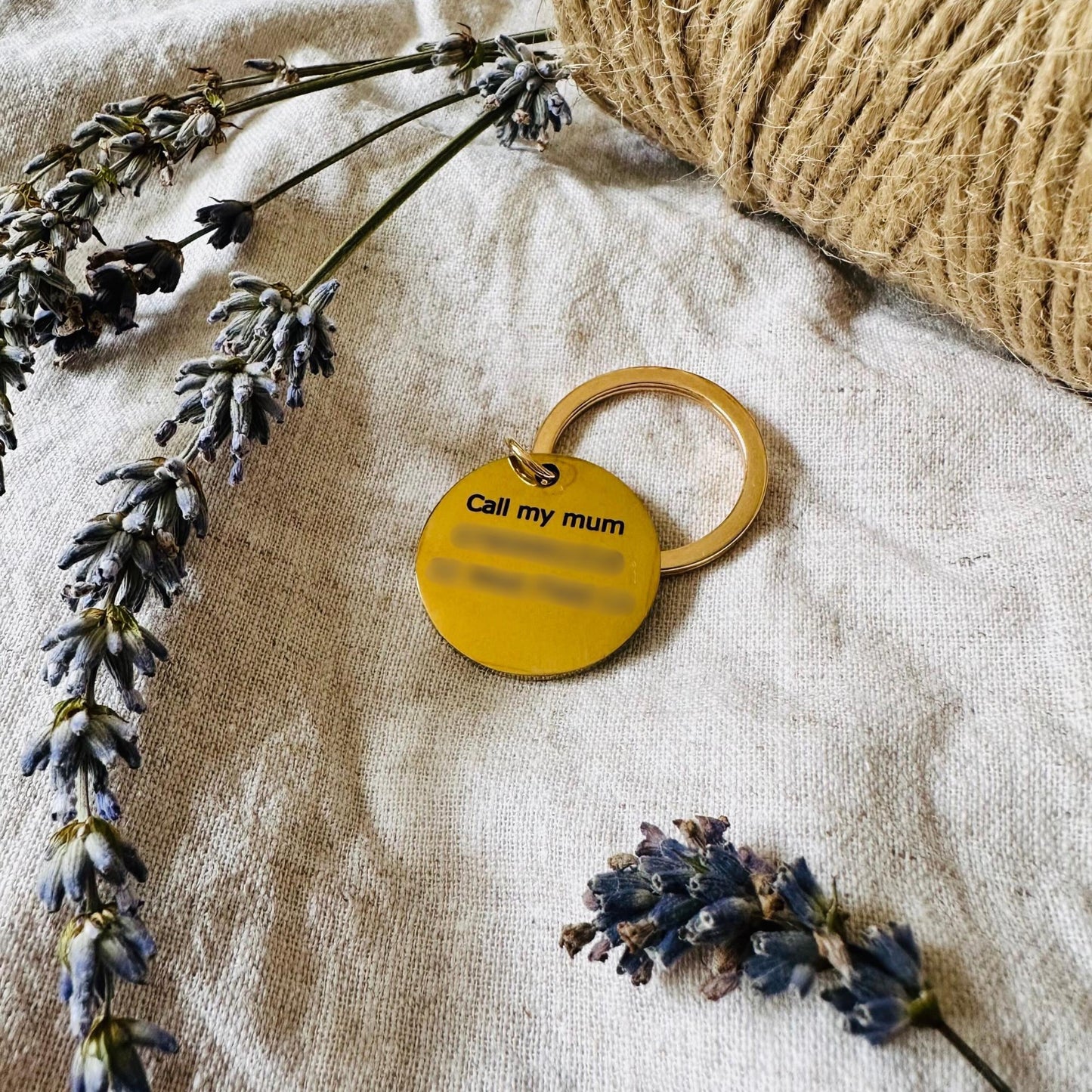 Gold dog tag with 'Call my mum' text on a textured surface with lavender flowers and twine.