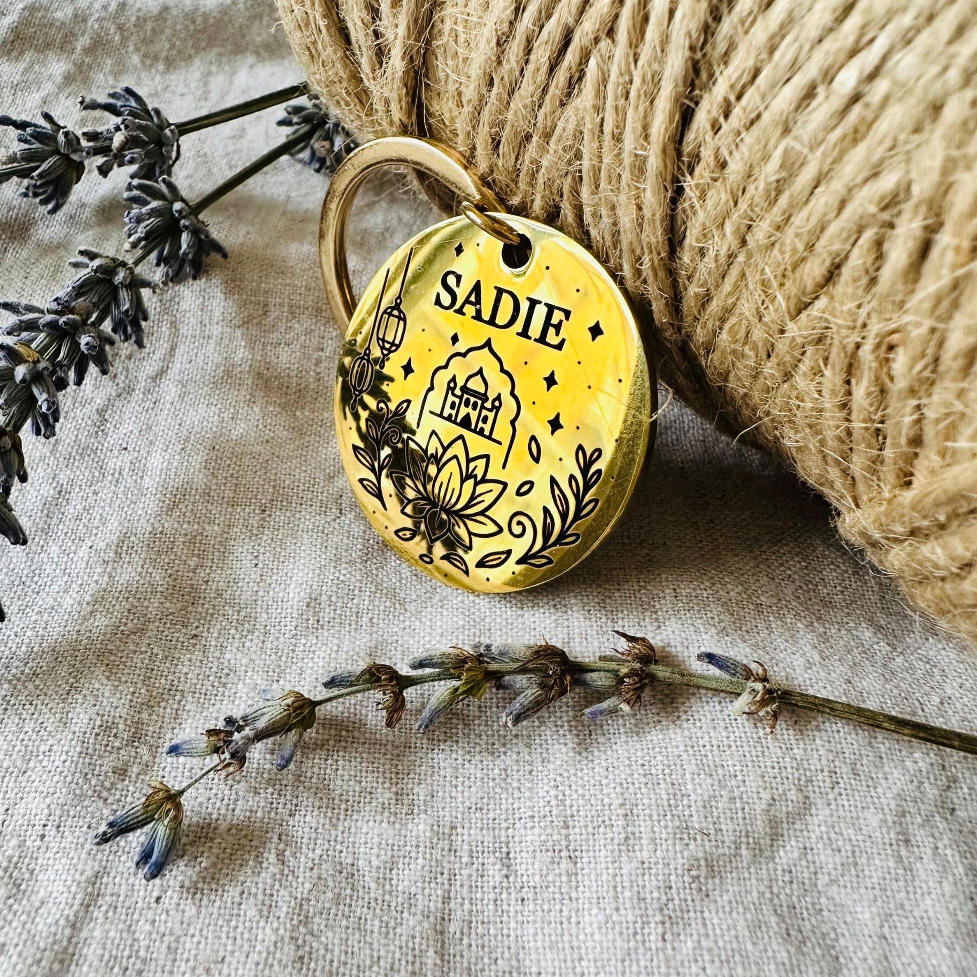 Gold pet tag with 'SADIE' engraved, surrounded by lavender and twine on a textured fabric background
