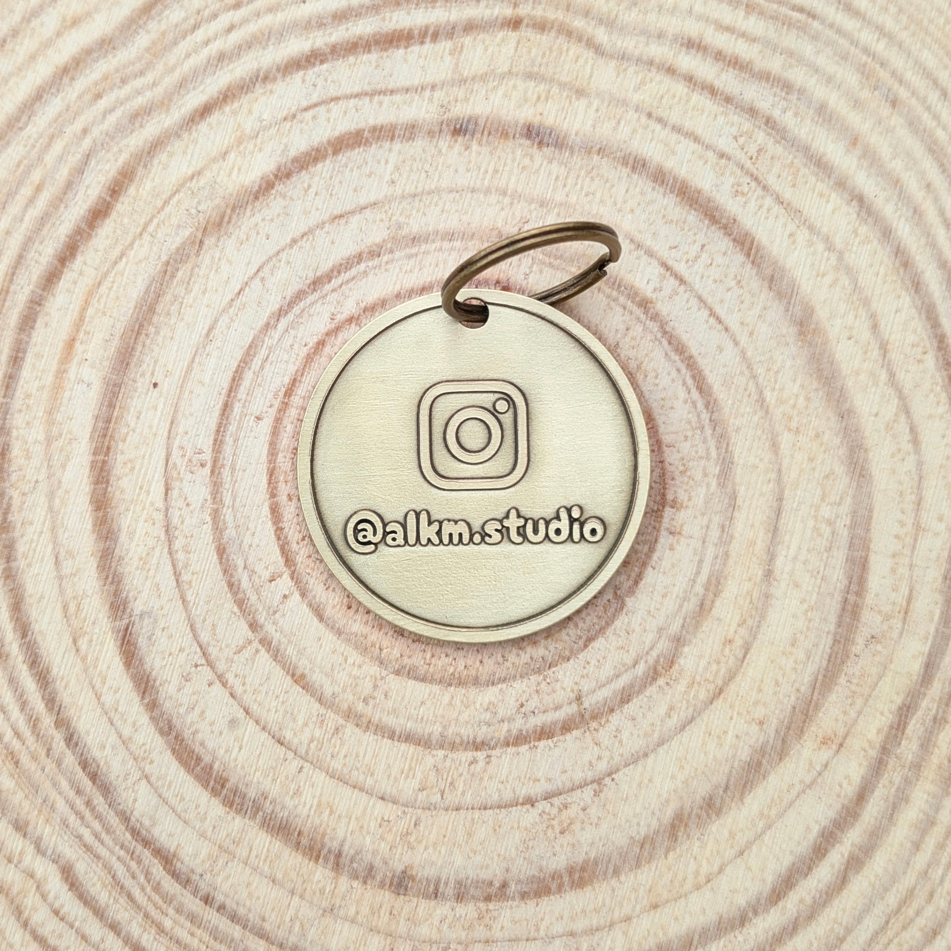 Bronze-colored round pendant with Instagram logo and '@alkm.studio' text on a wooden surface.