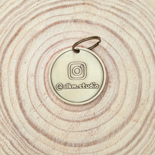 Bronze-colored round pendant with Instagram logo and '@alkm.studio' text on a wooden surface.