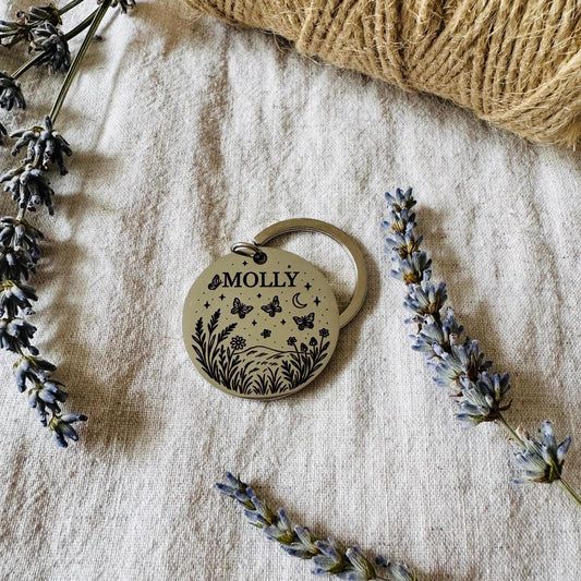 Round metal tag with 'MOLLY' engraved, surrounded by butterflies and flowers on a textured fabric background
