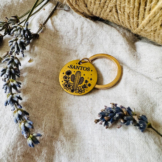 Gold dog tag with 'SANTOS' branding on a textured fabric background with lavender flowers.