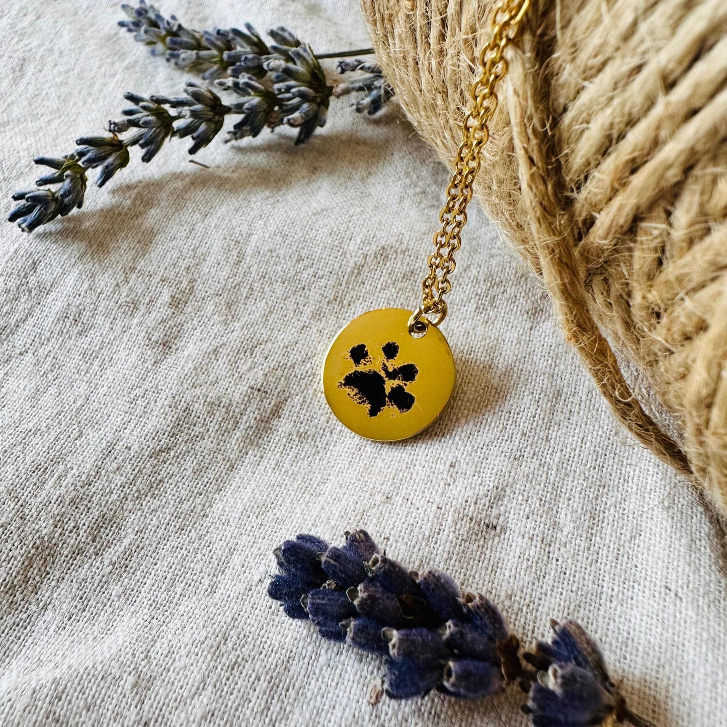 Gold necklace with a gold pendant featuring a black paw print, placed on a textured surface with lavender flowers.