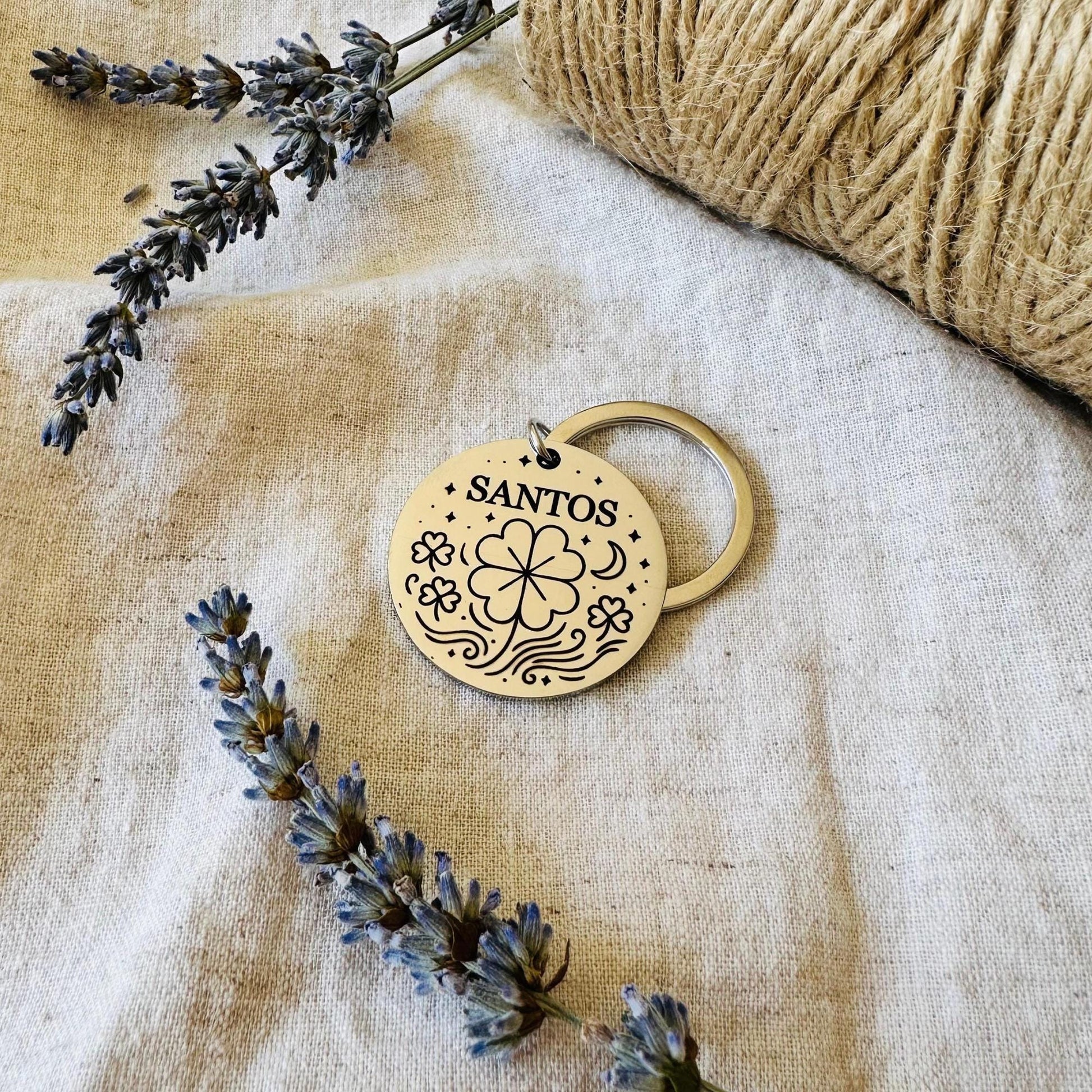 Dog Tag with engraved clover design and 'SANTOS' text, surrounded by lavender flowers on a textured fabric background.