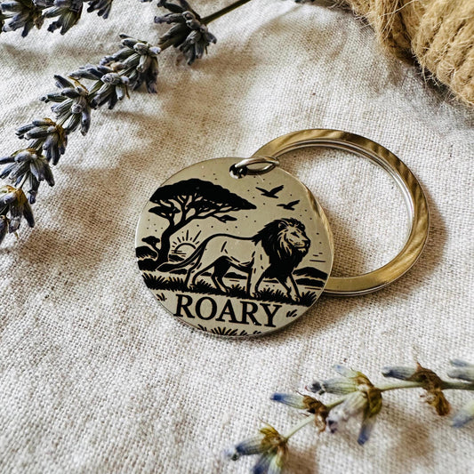 Dog tag with lion design and 'ROARY' text on a textured surface with lavender flowers.