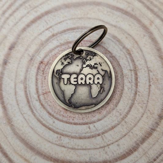 Brass Pet ID Tag with world map design and 'TERRA' text on a wooden surface