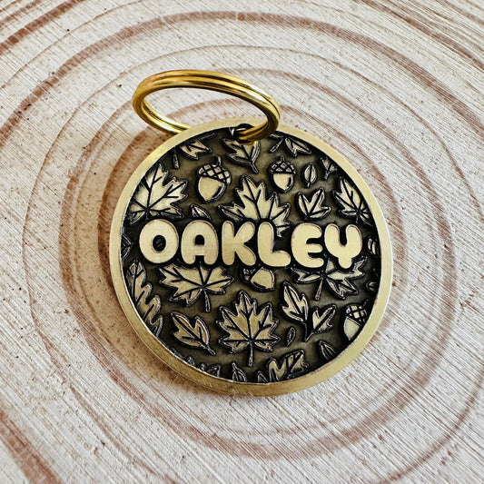 Deep engraved autumnal maple leaves and acorns on a brass id tag laying on a wooden surface.