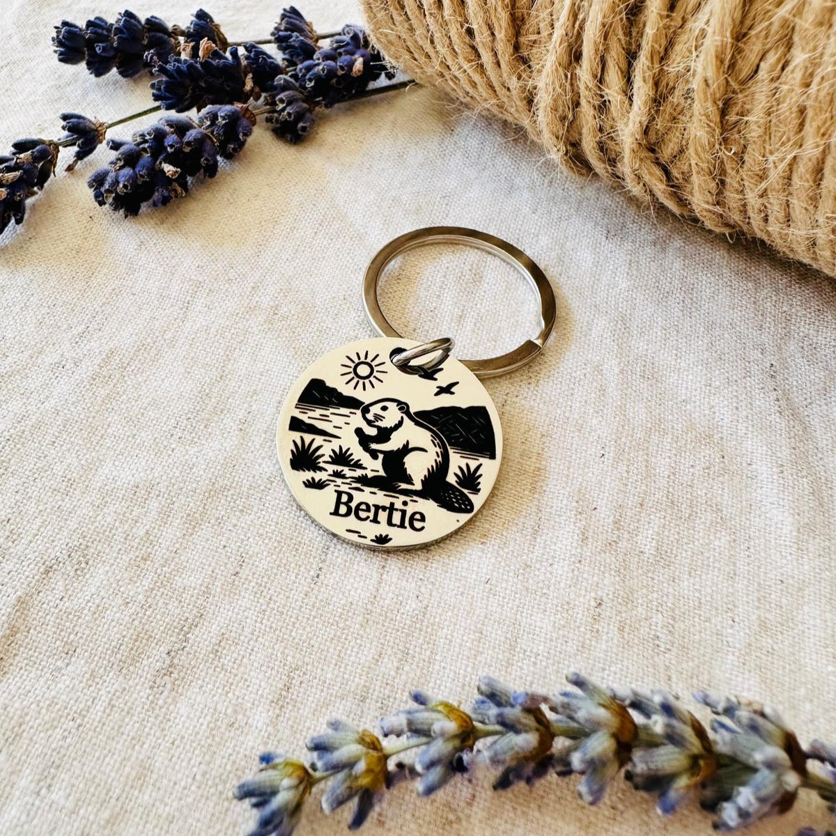 Pet id tag with beaver design and 'Bertie' engraving on a textured surface with lavender flowers.