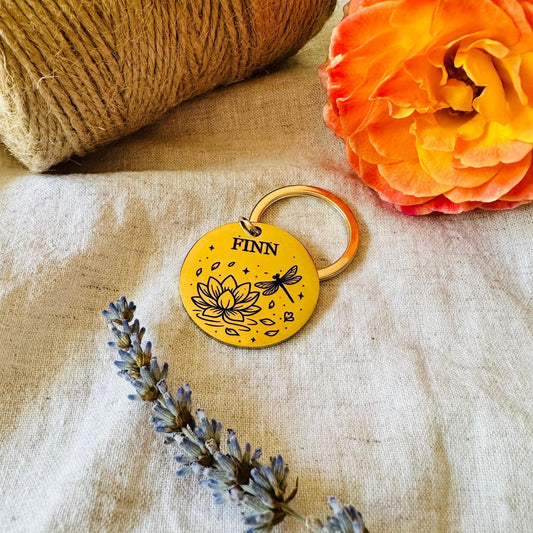 Gold tag with 'FINN' engraved, featuring a floral design, on a textured fabric background with flowers.