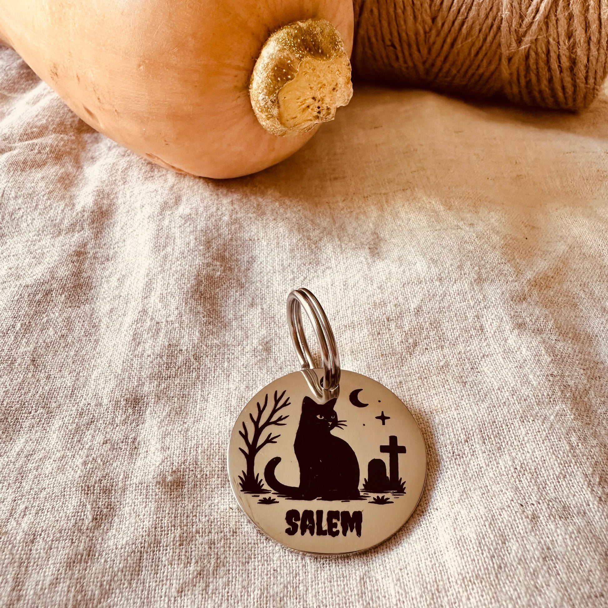 Dog tag with a cat silhouette and 'Salem' text on a rustic fabric background and a pumpkin.