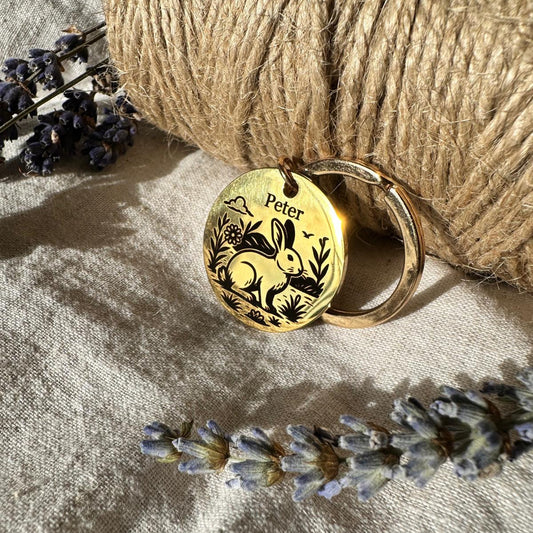 Gold pet id tag with engraved bunny and flower design on a textured surface with lavender flowers