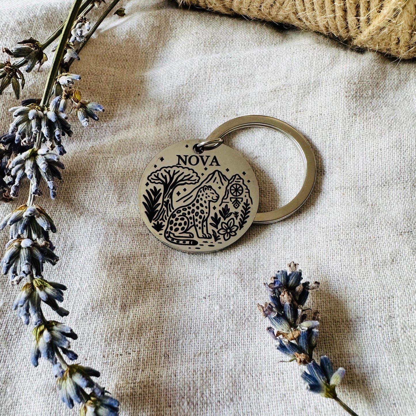 Round wooden keychain with 'NOVA' engraving on a textured fabric background with dried flowers.
