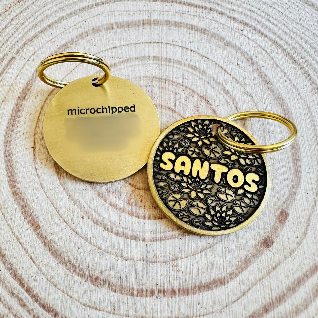 Brass dog tags one with deep engraved text another with lotus design on a textured wooden slab surface.