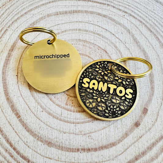 Brass dog tags one with deep engraved text another with lotus design on a textured wooden slab surface.