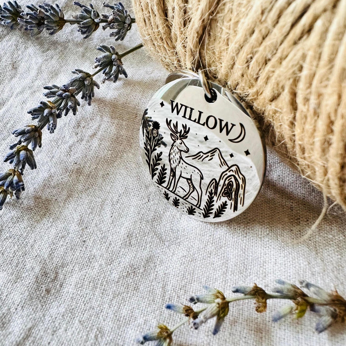 Silver dog tag with engraved design and 'Willow' text, surrounded by twine and dried flowers on a textured fabric background.