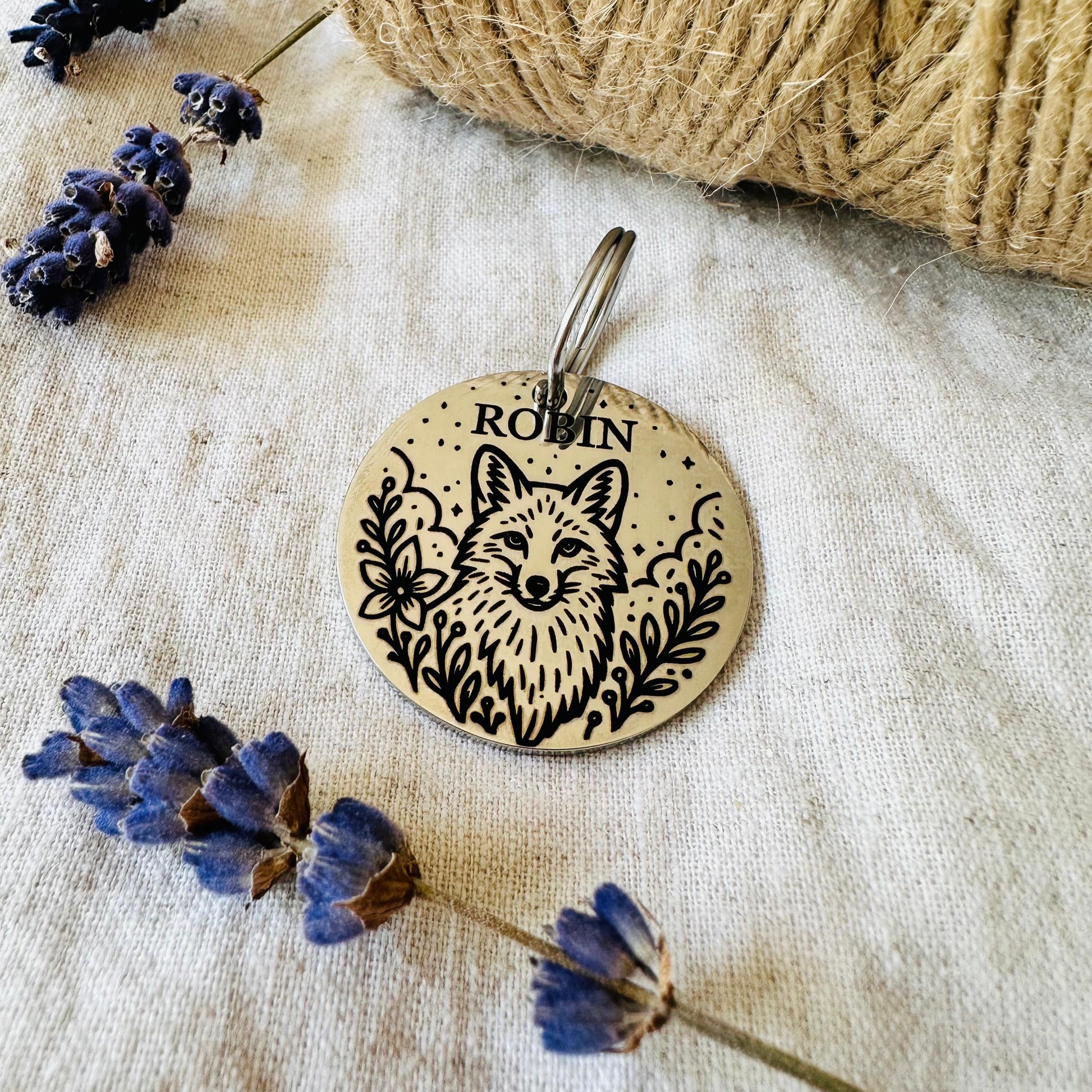 Round stainless steel pet id tag with fox design and 'Robin' engraving on a textured surface with lavender.