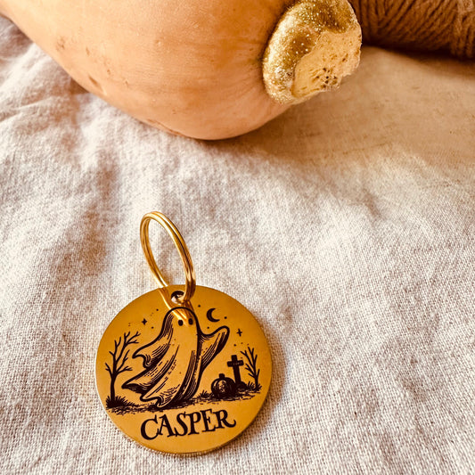 Gold pet ID tag with the name 'Casper' and a ghost engraving on a fabric background with a pumpkin and twine.