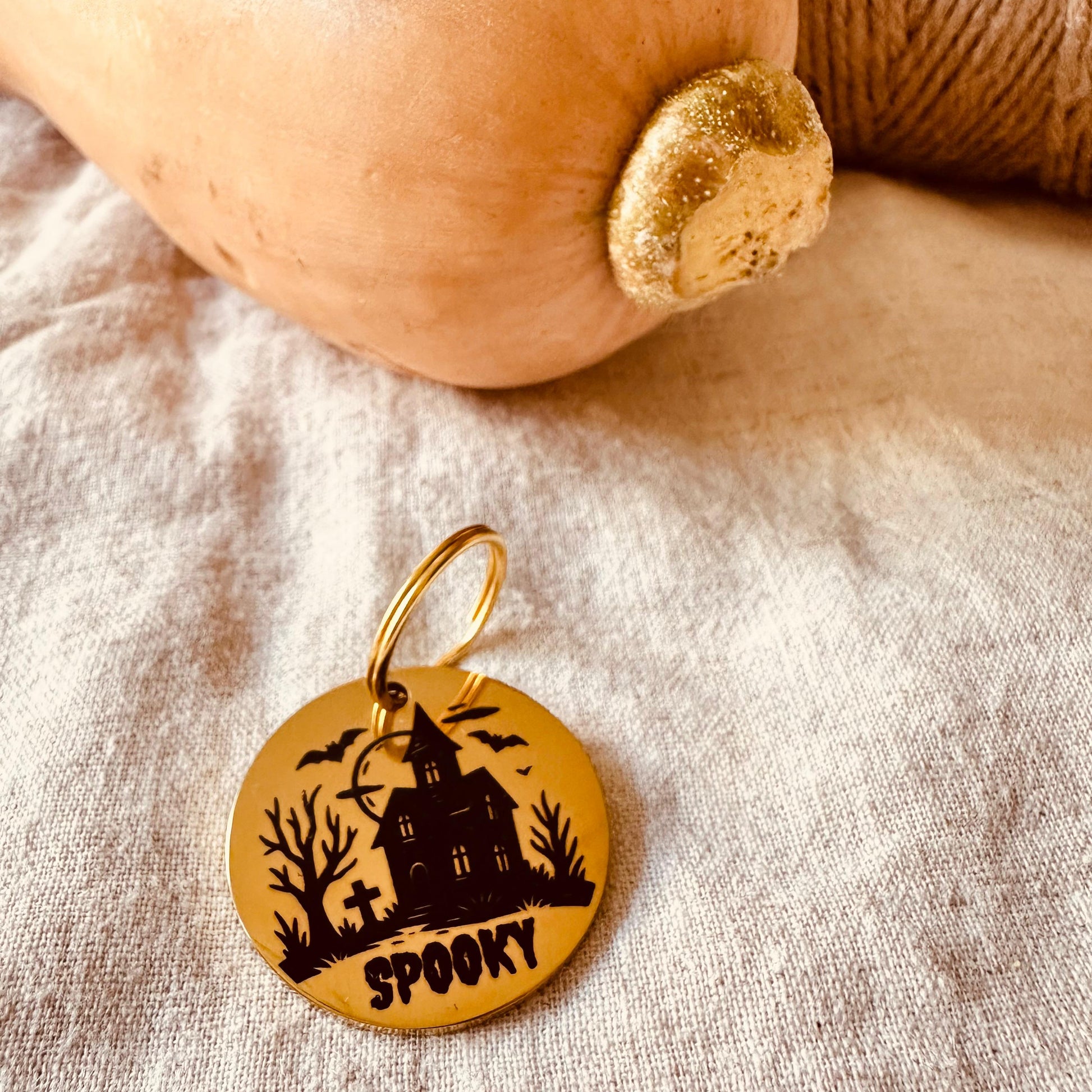 Gold pendant with the name 'Spooky' and haunted house design engraving on a fabric background with pumpkin and twine.