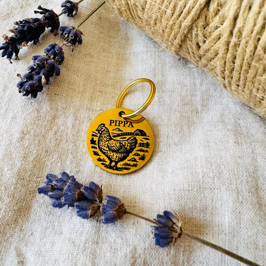 Yellow pet tag with a chicken design and 'Pippa' engraved, surrounded by lavender flowers and twine on a textured fabric background.