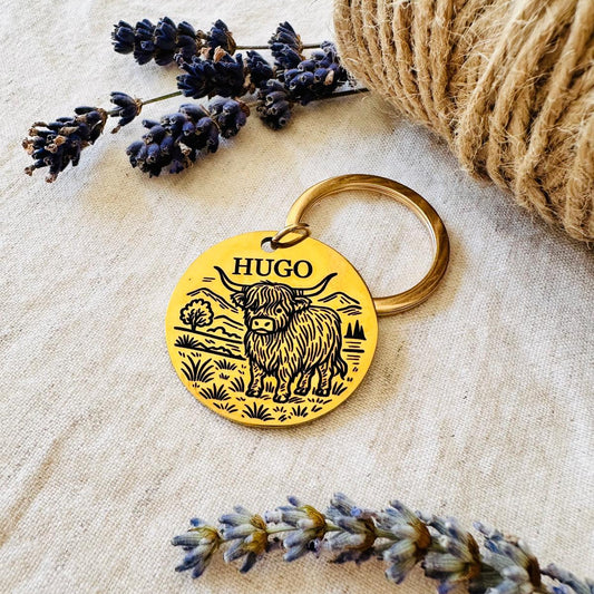Gold colour pet id tag with a highland cow design and 'Hugo' text, surrounded by lavender and twine, placed on a linen surface.