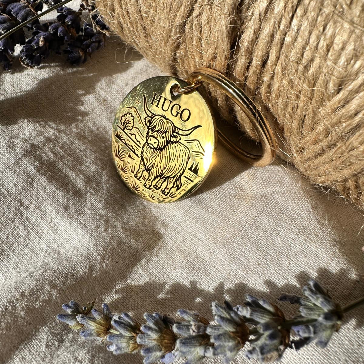 Gold pendant with 'HUGO' engraved, placed on a textured surface with lavender flowers.