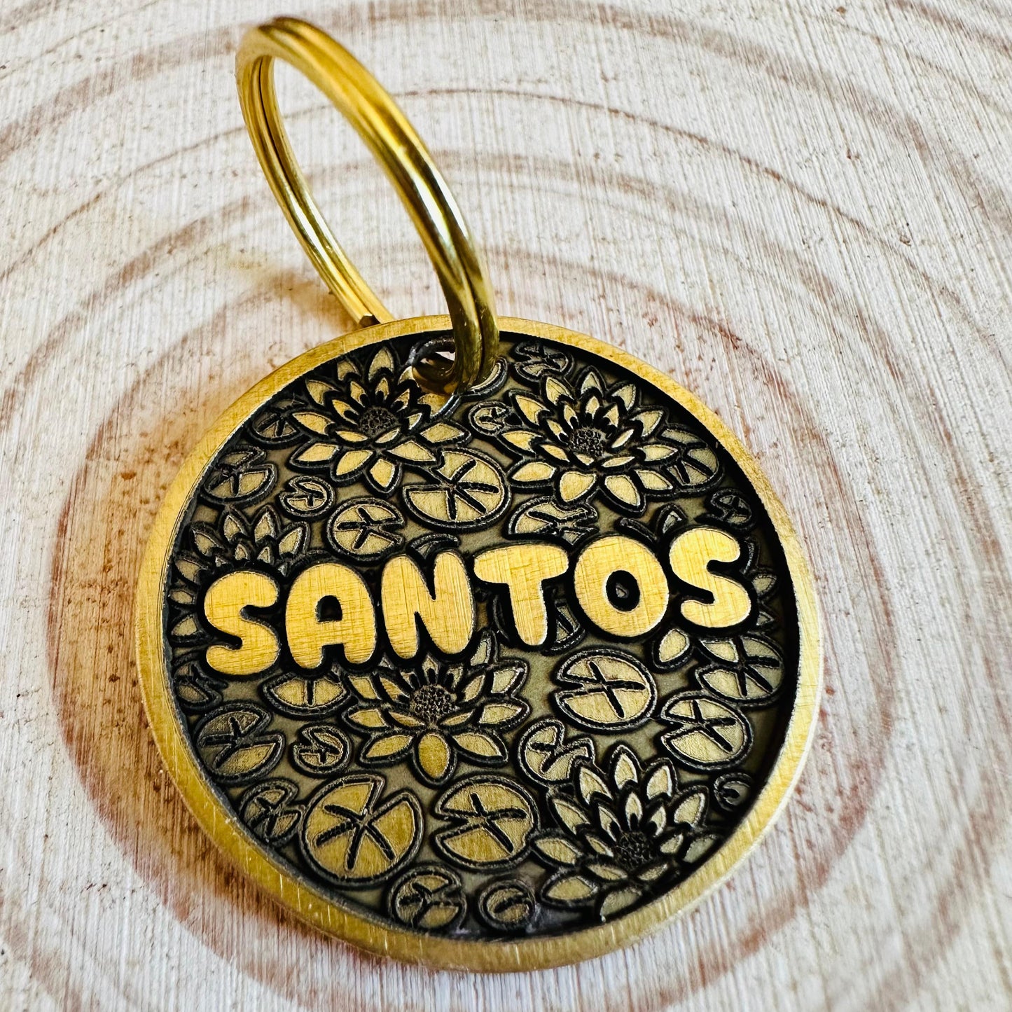 Gold and black pet tag with 'SANTOS' deep engraved on a floral lotus background laying on a wooden surface.