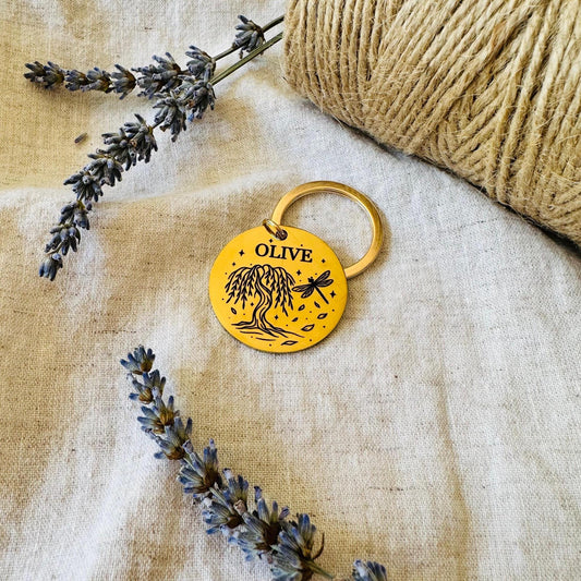 Gold dog tag with 'OLIVE' and willow tree design on a textured fabric background with lavender and twine.