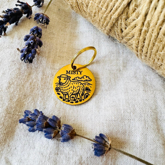 Gold finish stainless steel pet id tag with sheep design on a textured fabric background with lavender flowers
