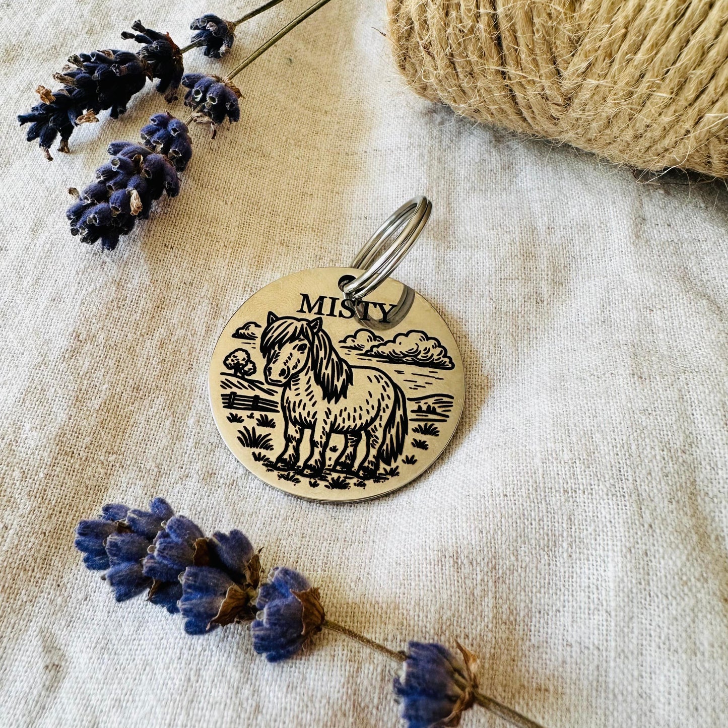 Round stainless steel pet id tag in silver with horse design on a textured fabric background with lavender flowers and twine.