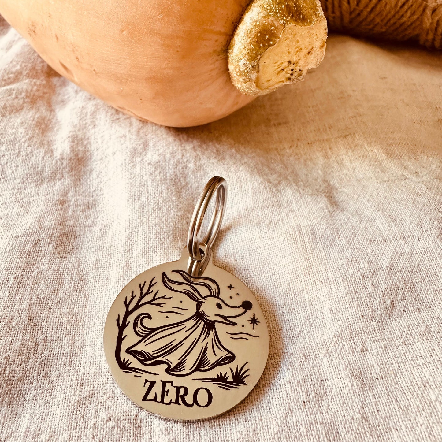 Stainless steel silver finish dog tag with engraved ghost dog design and 'ZERO' text on a fabric background with pumpkin and twine.