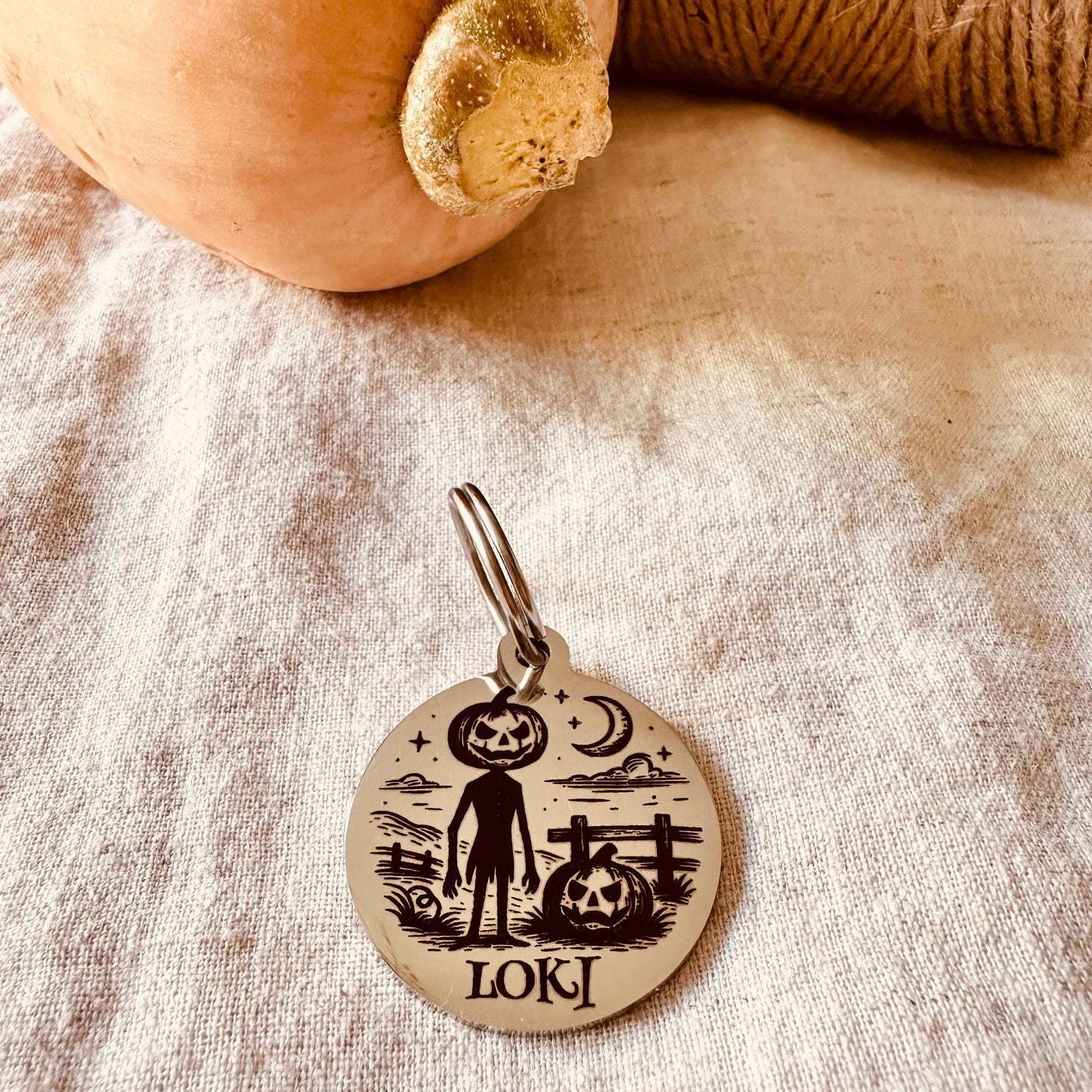 Silver finish pet tag with Jack O Lantern engraving and name Loki on a textured fabric background with a pumpkin and twine. 