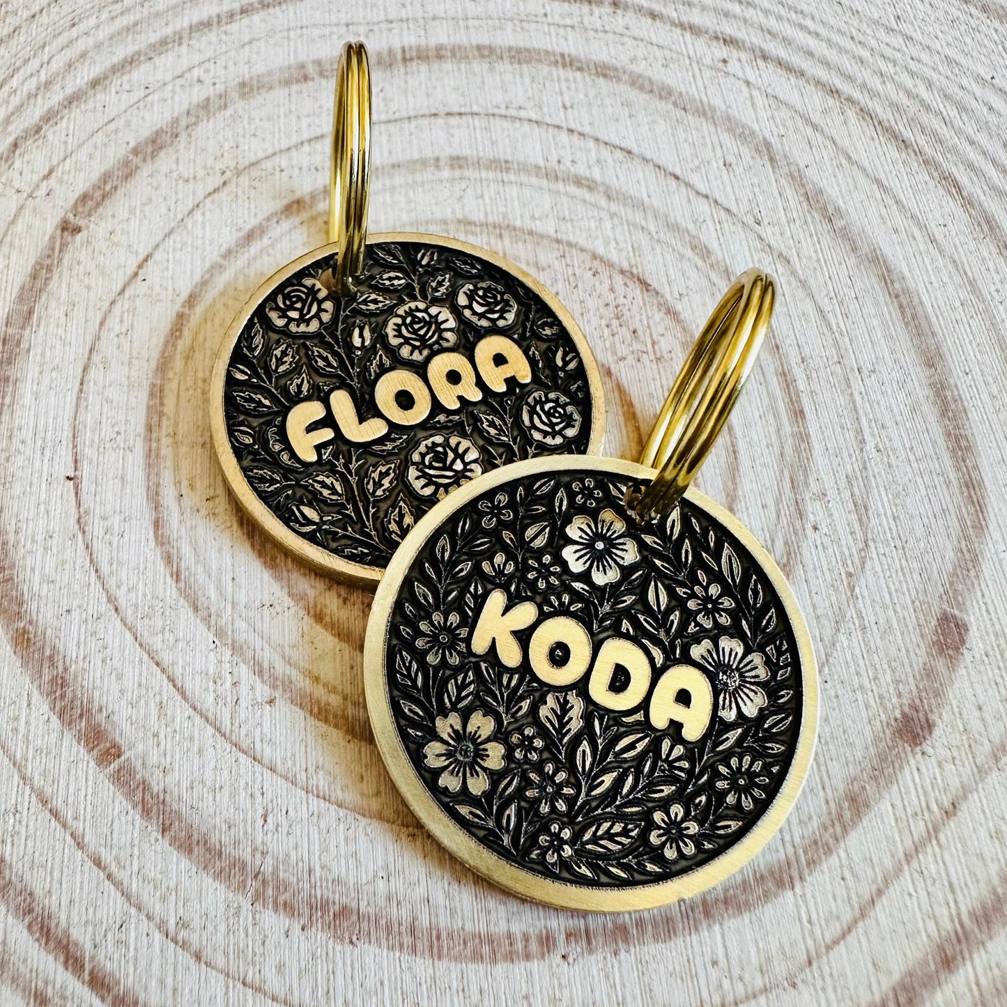 Two deep engraved black and gold dog tags with floral patterns on a wooden surface.