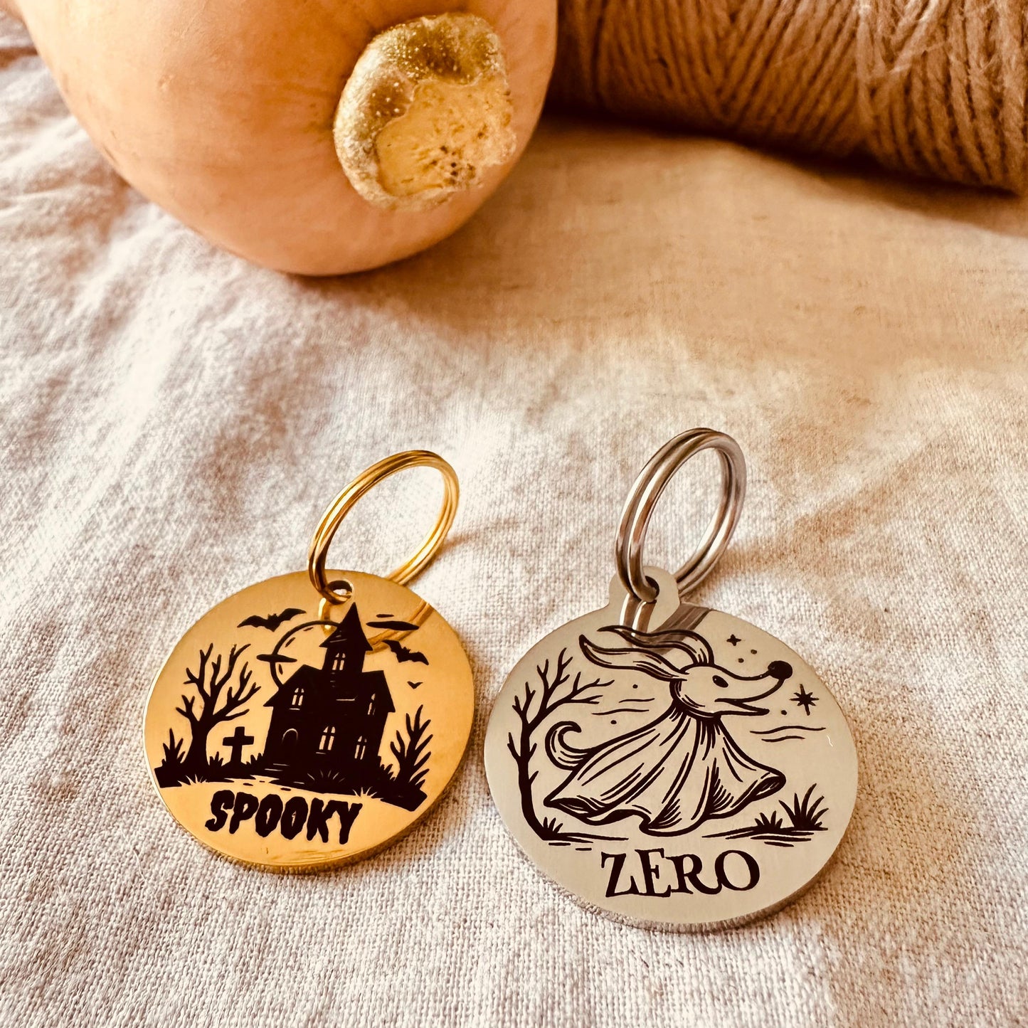 Two Halloween-themed dog tags on a textured surface with a pumpkin and yarn.