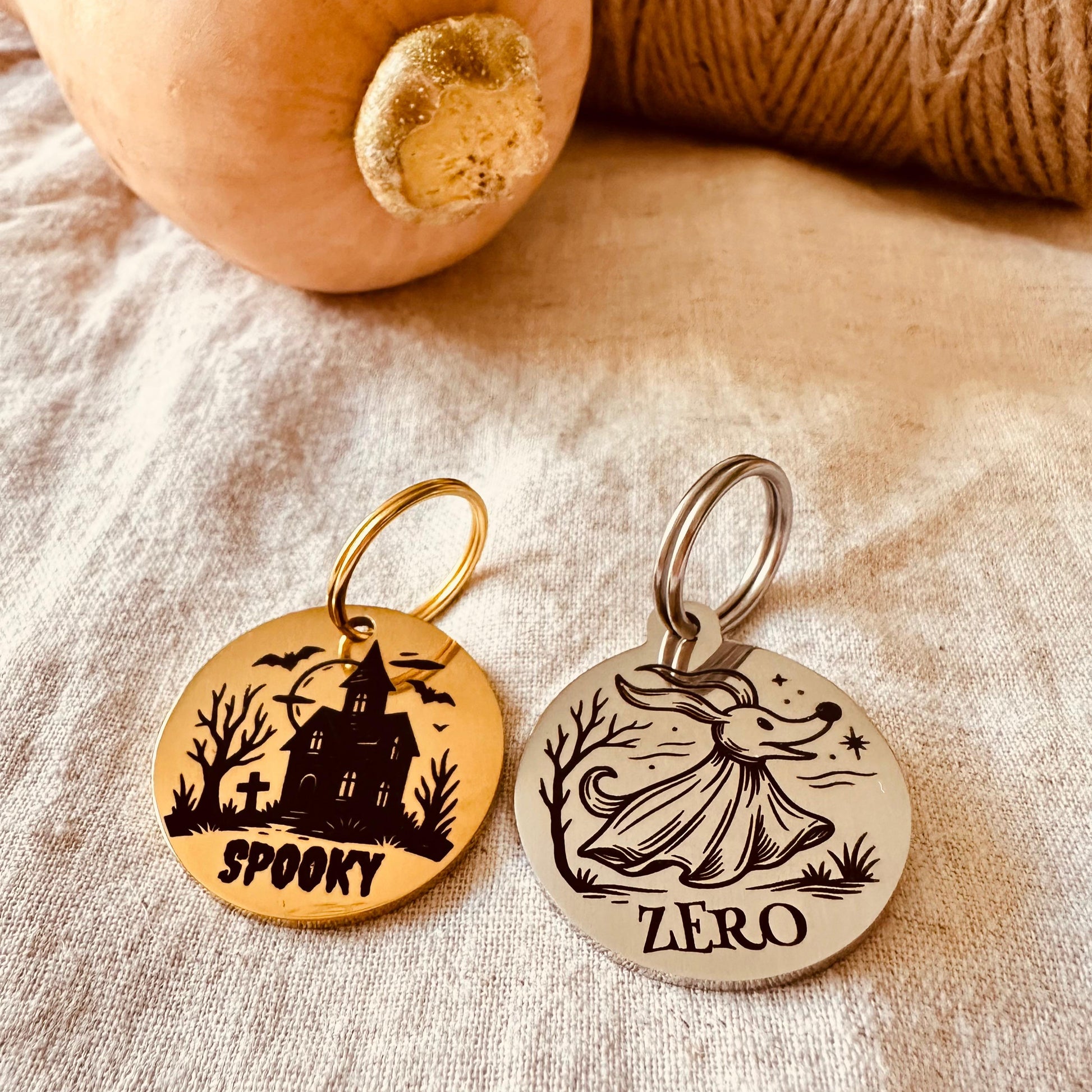 Two Halloween-themed dog tags on a textured surface with a pumpkin and yarn.