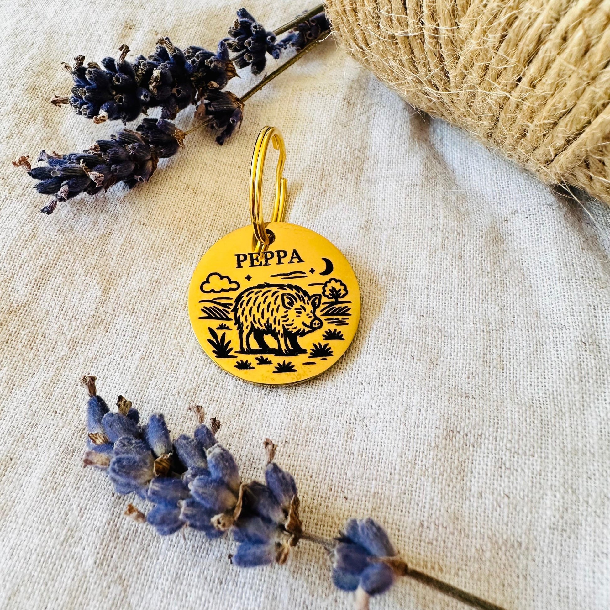 Gold pet tag with a boar design and 'Peppa' text, placed on a textured surface with lavender and twine.